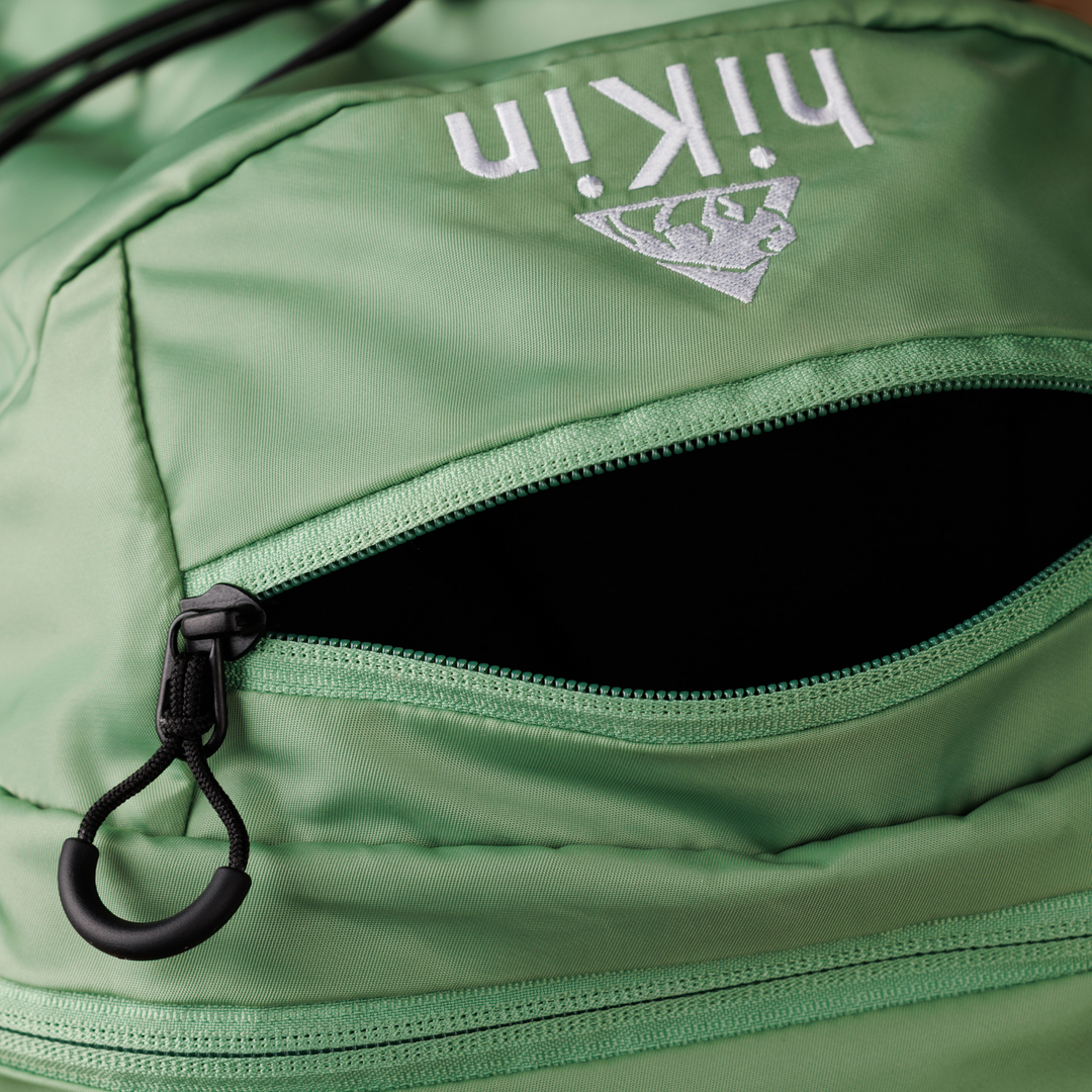 Close-up of a green backpack showing a zippered pocket with a black zipper pull and a white hikers logo on the fabric.