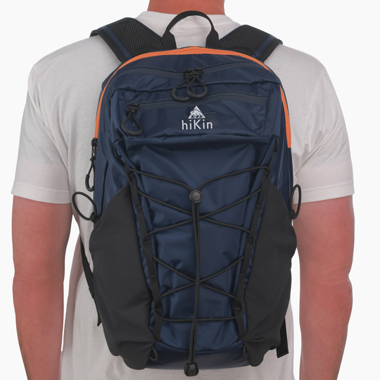 A person viewed from the back wearing a navy blue hikin backpack with black side pockets, black compression straps, and orange trim details.