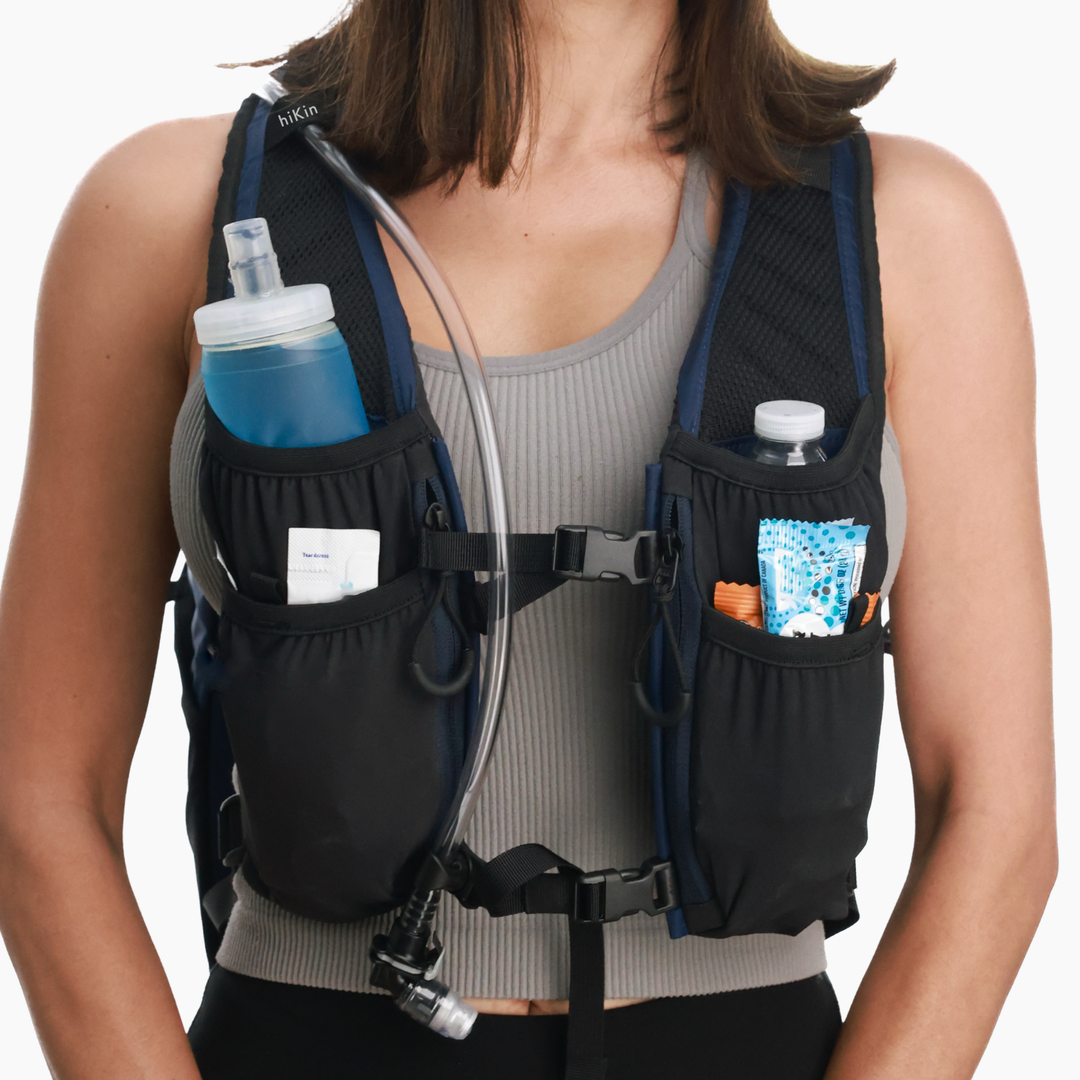 A close-up of a woman wearing a black hydration backpack with side pockets containing water bottles and a snack packet. The vest has a gray bib panel and black mesh straps, shown against a plain white background.