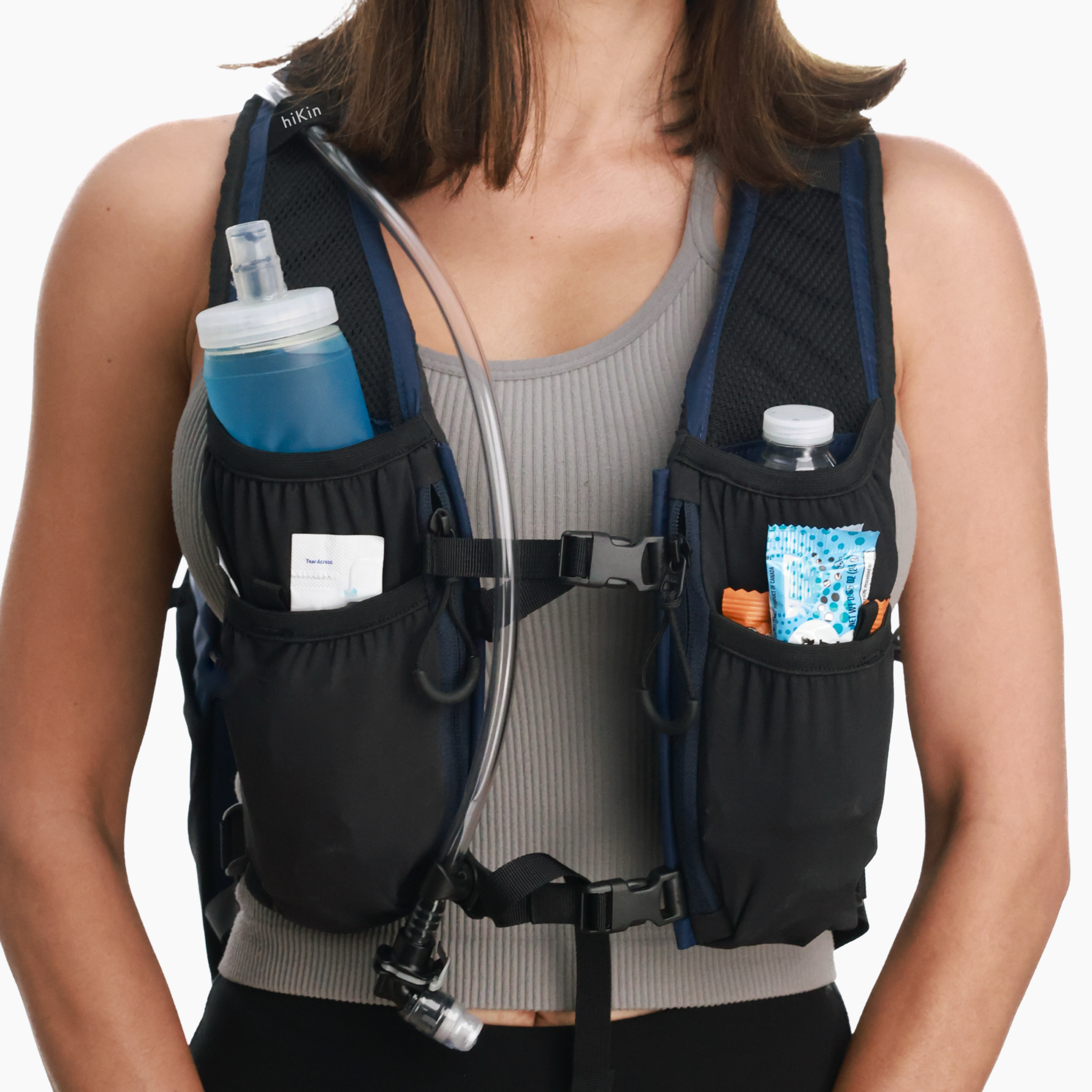 A close-up of a woman wearing a black hydration backpack with side pockets containing water bottles and a snack packet. The vest has a gray bib panel and black mesh straps, shown against a plain white background.