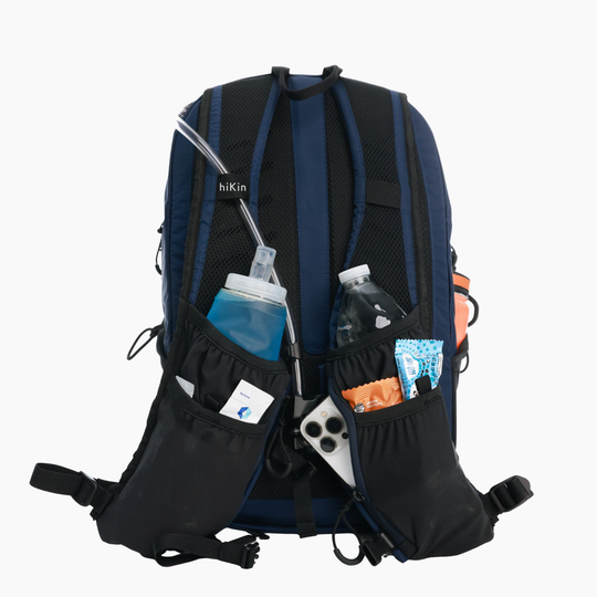 The back view of a hiking backpack in navy blue and black, shown with its side pockets filled with a water bottle, a phone, a snack packet, and a hydration tube, on a solid white background.