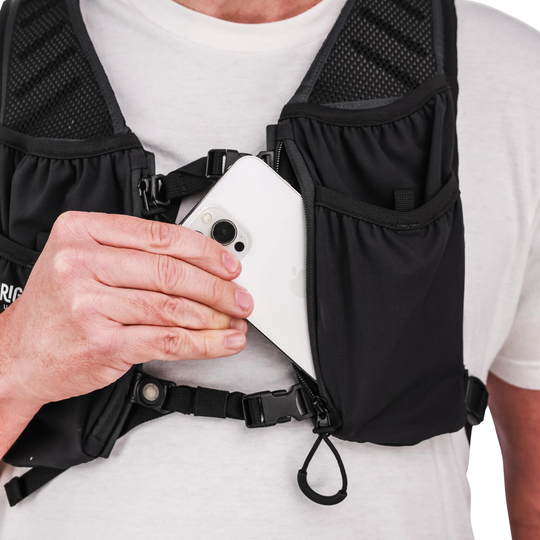 Close-up of a black running hydration vest worn by a person in a white t-shirt, with a hand holding a white smartphone inside the front pocket.