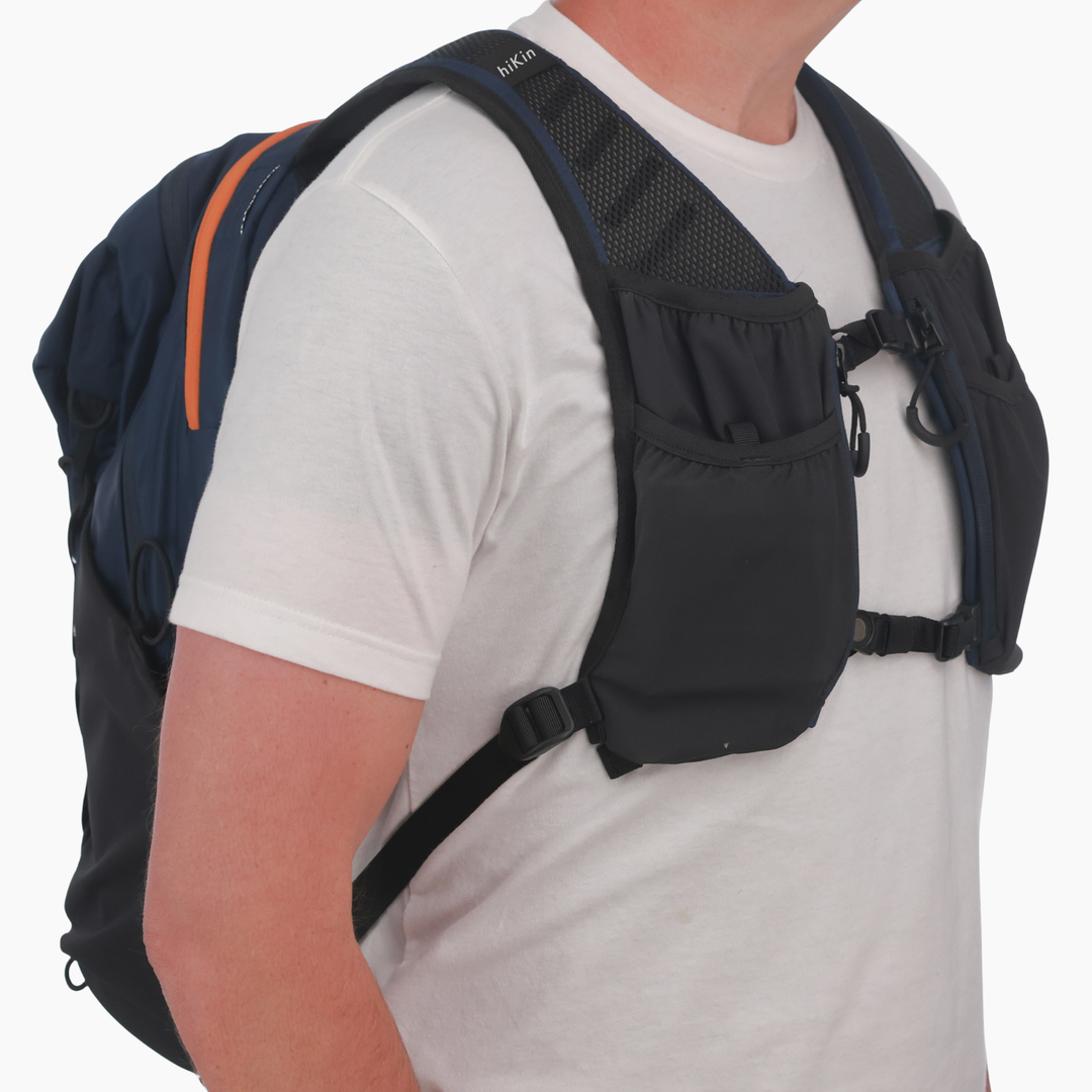 A close-up of a person wearing a black and navy blue hydration backpack with front pockets and shoulder straps over a white t-shirt, against a light gray background.