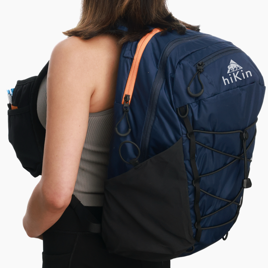 A woman from behind wearing a navy blue hikin backpack with black and orange accents, shown against a plain white background.
